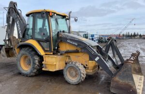 Retroescavadeira Volvo BL60B amarela e preta em canteiro de obras, vista lateral com cabine fechada e braço traseiro