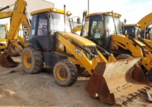Retroescavadeira JCB amarela com caçamba frontal, cabine fechada e braços hidráulicos em pátio de máquinas